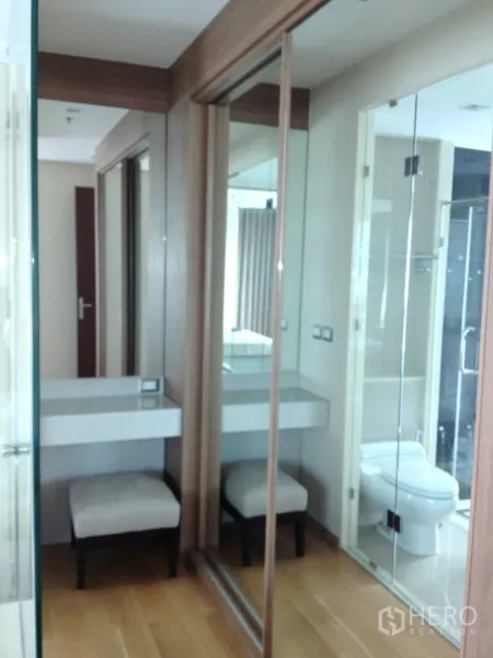 Condominium for rent in Ratchathewi, Bangkok - Dressing area with mirrored wardrobes, a vanity desk and access to the glass-partitioned bathroom.