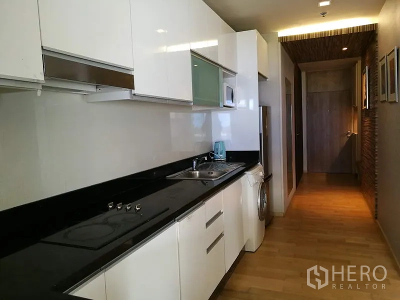 Condominium for rent in Watthana, Bangkok - Galley kitchen with glossy white cabinets, black countertop, cooktop, sink and in-unit washing machine.