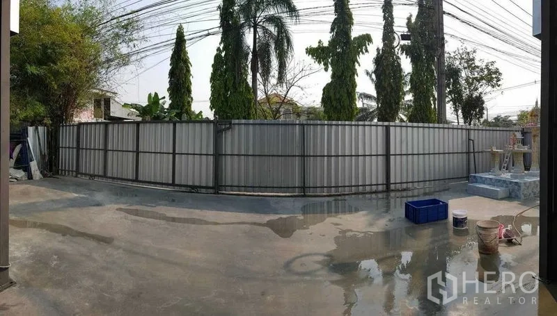 货栈 出租在Sai Mai, Bangkok - 地块周边金属围栏，外有棕榈树与架空电线，内有混凝土场地。