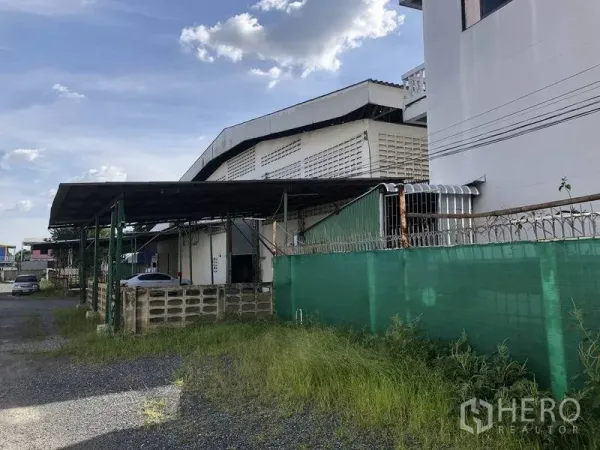 Warehouse for rent in Khlong Sam Wa, Bangkok - Warehouse exterior with covered loading bay, green perimeter fence and gravel driveway in Khlong Sam Wa.