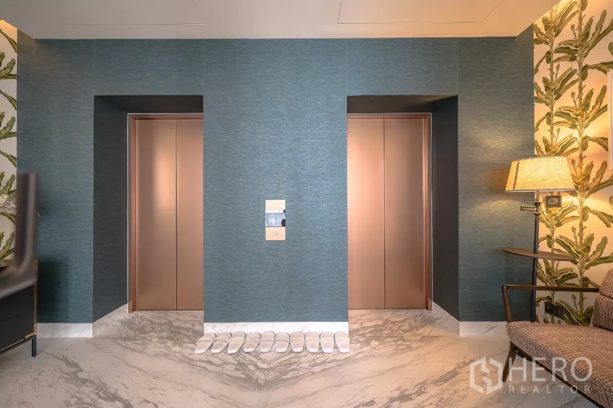 公寓 出租在Watthana, Bangkok - 对称双电梯的走廊，花纹墙面与整齐摆放的拖鞋。