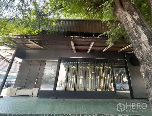零售空间 出租在Suan Luang, Bangkok - 单层零售店正立面，大面积展示窗，树荫与金属雨棚覆盖。