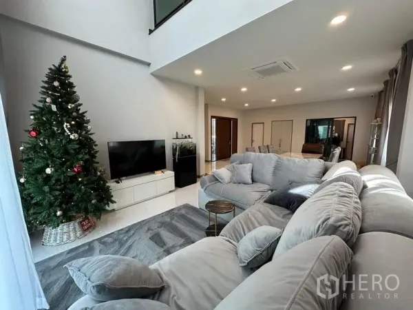 独立式住宅 出租在Bang Phli, Samut Prakan - 挑高客厅，L 型沙发、电视柜与落地窗帘旁的装饰树。