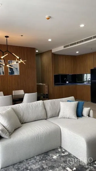 Condominium for rent in Pathum Wan, Bangkok - Open-plan living with light gray sofa, wood accents and sleek black-gloss kitchen.
