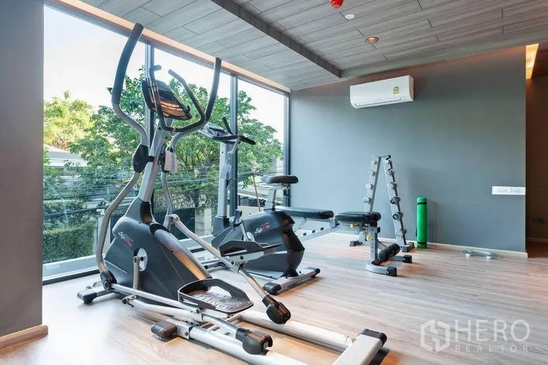 Condominium for rent or sale in Watthana, Bangkok - Light-filled fitness room with elliptical machines, exercise bike, free weights and floor-to-ceiling windows overlooking greenery.