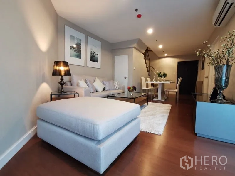 Condominium for rent or sale in Huai Khwang, Bangkok - Spacious open-plan living and dining room with staircase and large ottoman.