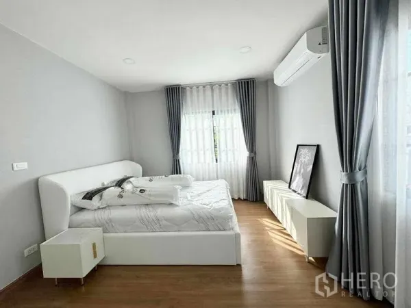 Detached House for rent or sale in Bang Phli, Samut Prakan - Airy bedroom with white bed, wood flooring, curtains, wall-mounted air conditioner and low cabinet.