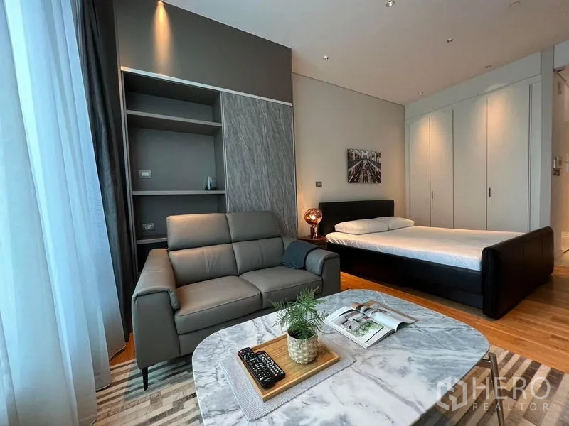 Condominium for rent in Pathum Wan, Bangkok - Open‑plan studio showing grey sofa, marble table, bed and full‑height wardrobes on wood flooring