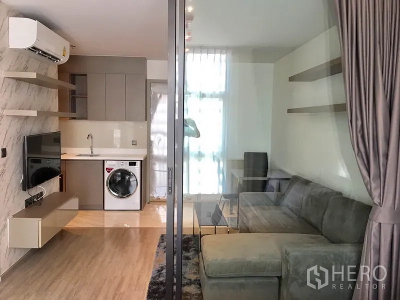 Condominium for rent in Watthana, Bangkok - Compact kitchen with a washing machine and wall-mounted TV opposite a glass-partitioned lounge.