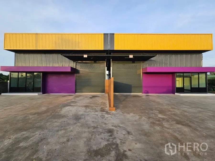 คลังสินค้า ให้เช่าในLam Luk Ka, Pathum Thani - มุมมองด้านหน้าของโกดังสองยูนิตสมัยใหม่ หลังคาเหลือง กันสาดสีม่วง ประตูม้วนคู่ พร้อมลานคอนกรีตกว้างในลำลูกกา