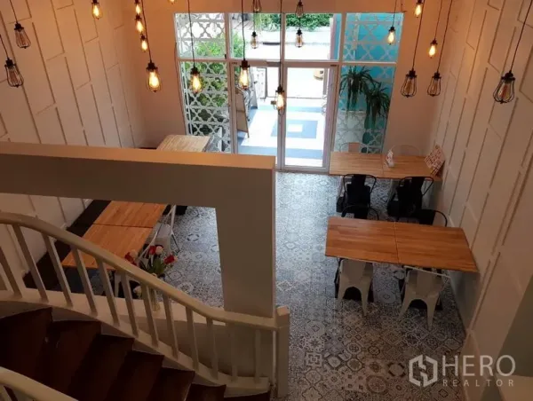 零售空间 出租在Bang Rak, Bangkok - 双层挑高的商铺内部，吊灯、木桌、花纹地砖与玻璃门，从楼梯俯视。