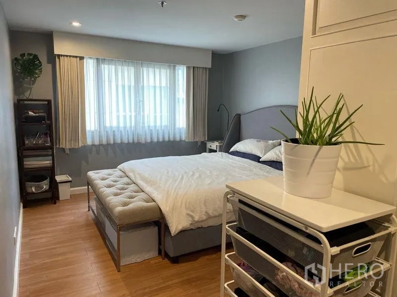 Condominium for sale in Watthana, Bangkok - Spacious master bedroom with large bed, bench, wood floors and plant by the window.