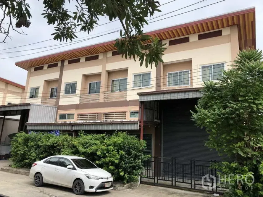 联排别墅 出售在Bang Bua Thong, Nonthaburi - 三层商业联排外立面，带院落围栏与卷帘门，绿化环绕。