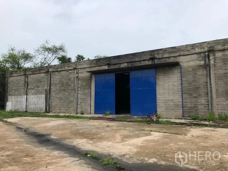 货栈 出租在Pak Kret, Nonthaburi - 单层仓库外观，灰色砌块墙与宽大的蓝色大门，带装卸空地。