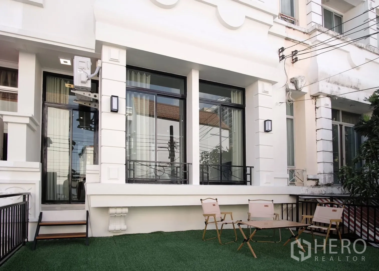 Townhouse for rent in Bang Phli, Samut Prakan - Townhouse facade with tall black-framed windows and a small artificial grass patio with outdoor chairs.