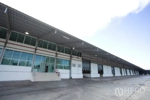 货栈 出租在Si Racha, Chon Buri - 现代仓库外立面，长条有顶装卸月台，玻璃办公区与多扇卷闸门。