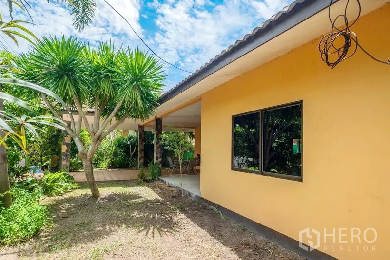 Detached House for sale in Bang Lamung, Chon Buri - Side garden with mature trees beside the home’s yellow exterior and patio.