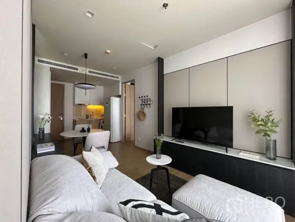 Condominium for rent in Watthana, Bangkok - TV wall with large flat screen, light gray sectional sofa and small round tables facing the kitchen entrance.
