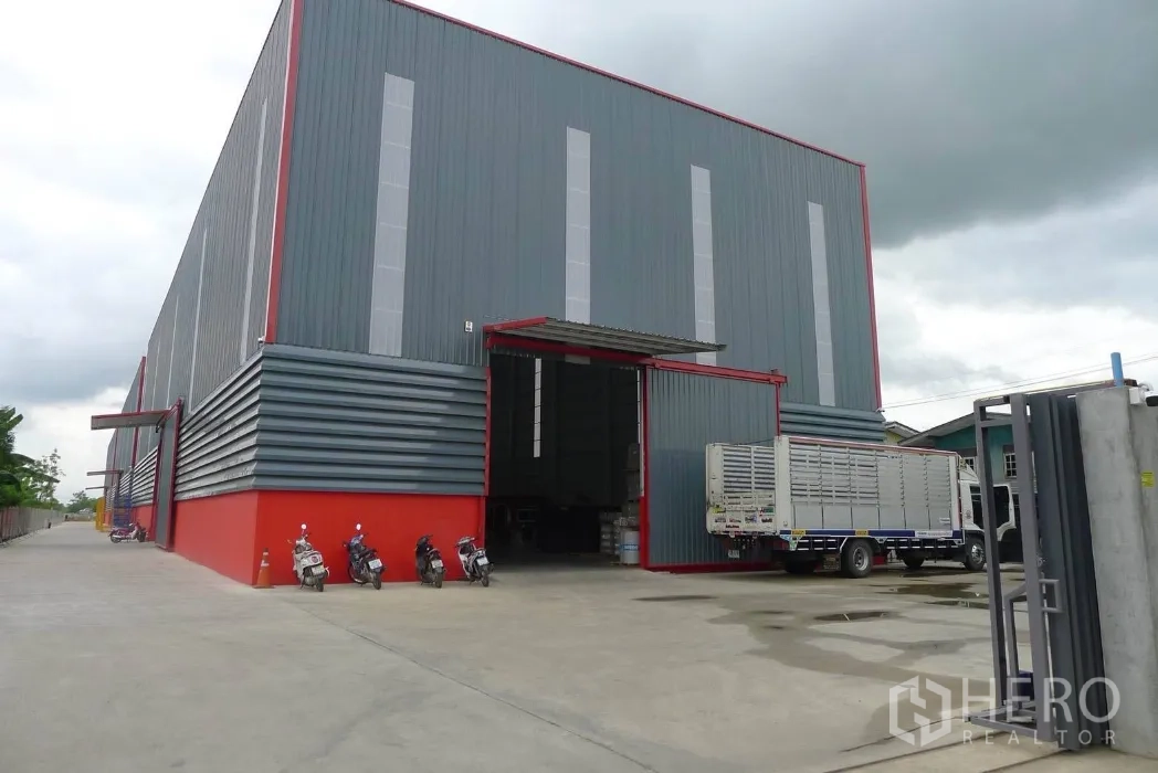 Warehouse for rent in Khlong Luang, Pathum Thani - Modern high-clearance warehouse with grey metal facade, red trim, large sliding loading door and wide concrete yard for trucks.