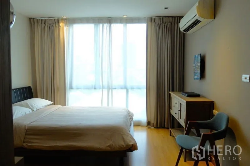 Condominium for sale in Watthana, Bangkok - Bedroom with full-height windows, air conditioner and a small working corner with chair.