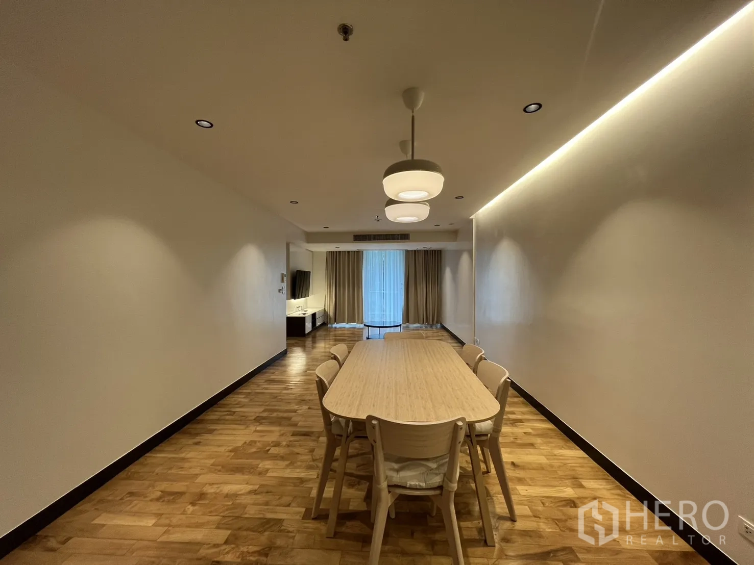 Apartment for rent in Watthana, Bangkok - Open-plan dining and living room with wood floors, pendant lights, a long table, media console, and sliding glass doors with curtains.