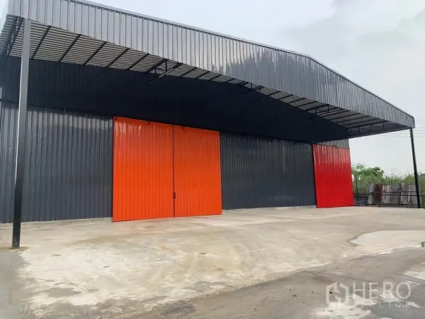 货栈 出租在Khan Na Yao, Bangkok - 黑色金属仓库外立面，橙色大推拉门与带顶装卸区，位于曼谷康那耀。