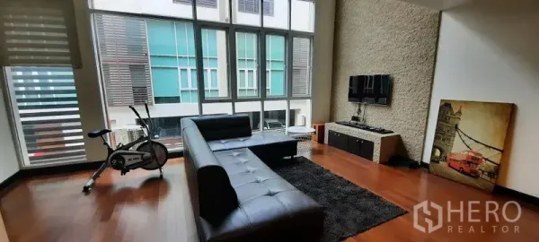 ทาวน์เฮาส์ ให้เช่าหรือขายในKhlong Toei, Bangkok - ห้องนั่งเล่นเพดานสูง หน้าต่างสูงจรดพื้น โซฟาหนังสีดำ ผนังทีวี และจักรยานออกกำลังกาย