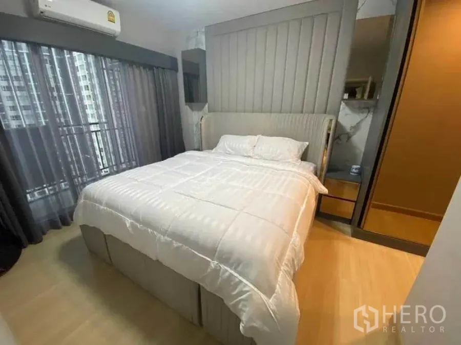 คอนโด ให้เช่าหรือขายในHuai Khwang, Bangkok - ห้องนอนคอนโดสไตล์โมเดิร์นในห้วยขวาง มีหัวเตียงบุผ้า ชุดเครื่องนอนสีขาว ตู้เสื้อผ้าบานกระจก แอร์ และหน้าต่างสูงชนฝ้าเห็นวิวเมือง