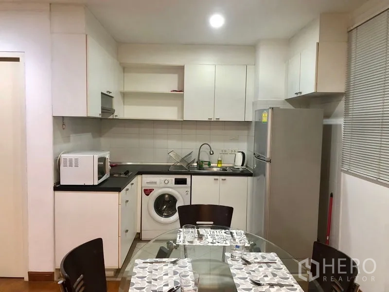 Condominium for rent or sale in Watthana, Bangkok - Compact kitchen with white cabinets, sink, fridge, microwave, and washing machine next to the dining area.