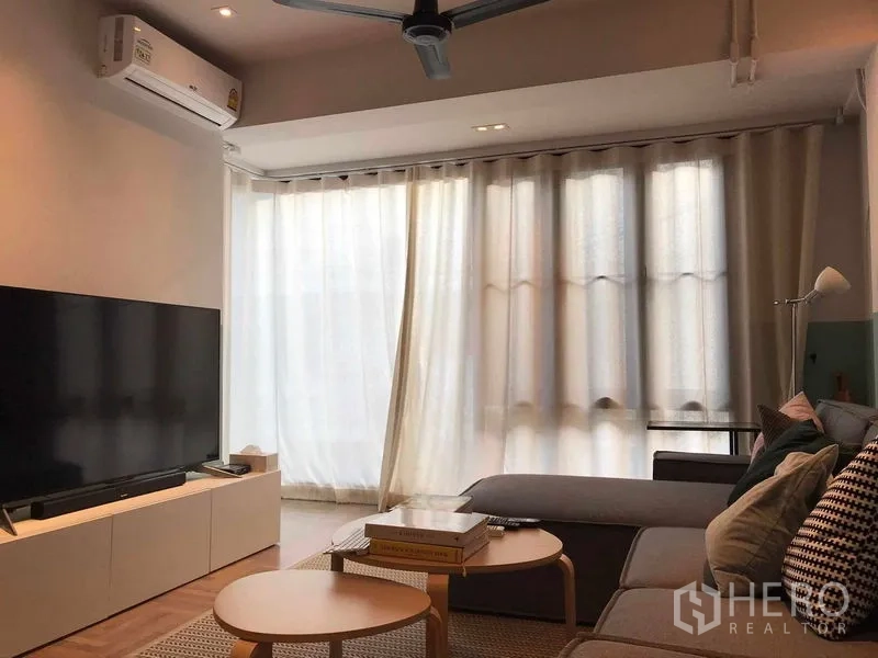 Townhouse for rent or sale in Khlong Toei, Bangkok - Bright living room with sheer curtains, ceiling fan, L‑shaped sofa and TV console.