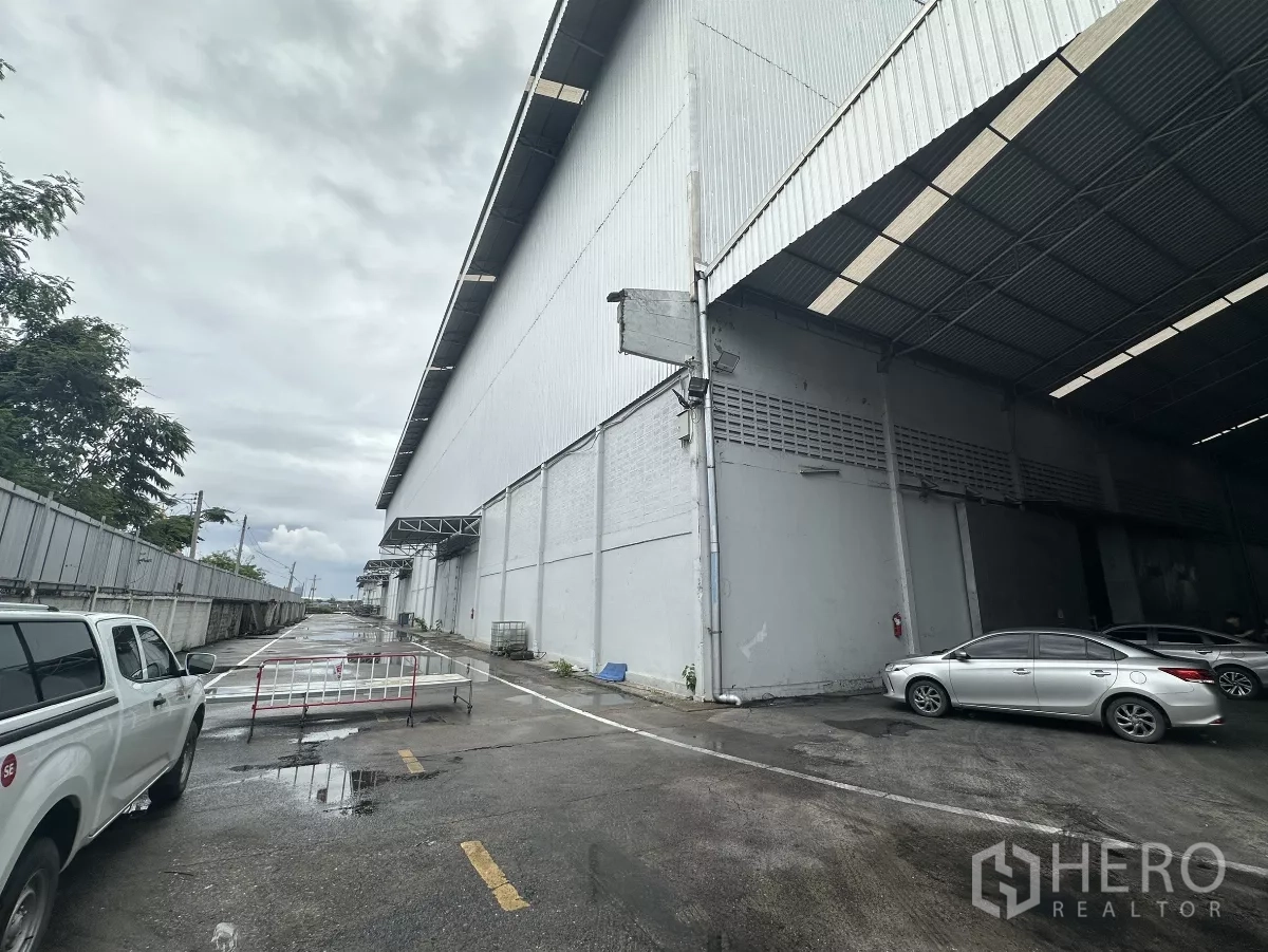 货栈 出租在Mueang Pathum Thani, Pathum Thani - 仓库外立面与带顶装卸区，宽阔车道及现场停车位（巴吞他尼）。