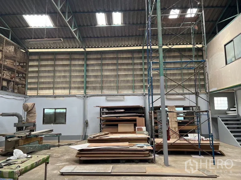货栈 出租在Khlong Sam Wa, Bangkok - 开阔生产空间，带高脚脚手架、通风百叶墙与堆放板材的区域，屋顶有天窗。