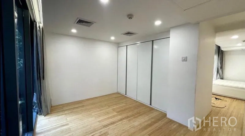 Condominium for rent in Watthana, Bangkok - Bright bedroom with a wall of white wardrobes, large window, and wood floors.