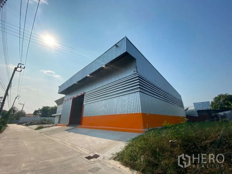 Warehouse for rent in Don Mueang, Bangkok - Corner angle of a steel warehouse with an orange base and ramp access on a quiet concrete road.