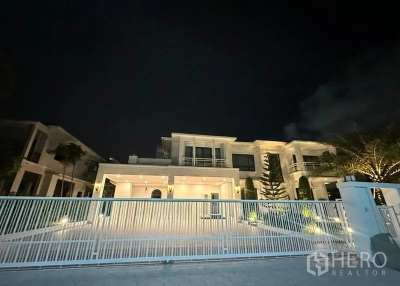 Detached House for rent in Bang Phli, Samut Prakan - Night view of the illuminated modern house behind a white metal gate.