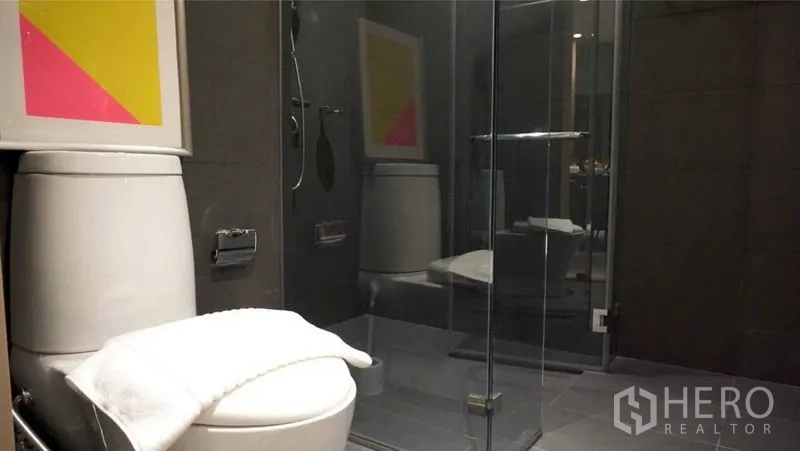 Condominium for rent in Pathum Wan, Bangkok - Modern bathroom with glass shower enclosure, wall fixtures and a toilet topped with a folded towel.