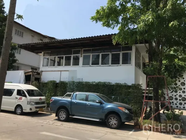 พื้นที่ขายปลีก ให้เช่าในWatthana, Bangkok - อาคารค้าปลีกสีขาวสองชั้น หน้าต่างยาว แนวรั้วต้นไม้ และที่จอดรถริมถนนใต้ร่มไม้