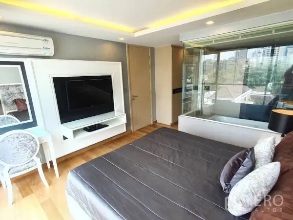 Condominium for rent in Watthana, Bangkok - Bedroom with built-in TV wall, desk, and glass-walled bathtub overlooking greenery and city views.
