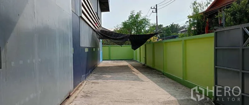 Warehouse for rent in Mueang Samut Sakhon, Samut Sakhon - Side service lane beside metal-clad warehouse with green boundary wall and concrete paving.