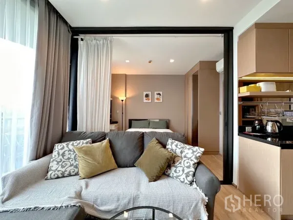 Condominium for sale in Watthana, Bangkok - Cozy living room with gray sofa, sliding glass partition opening to the bedroom and sheer curtains.