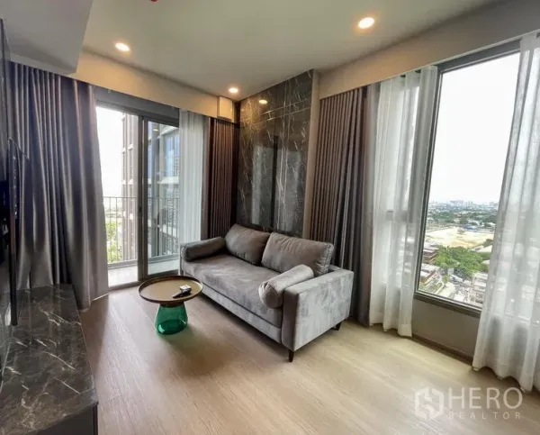 Condominium for rent in Samut Prakan - Modern living room with gray sofa, marble TV wall, floor-to-ceiling windows and a balcony city view.