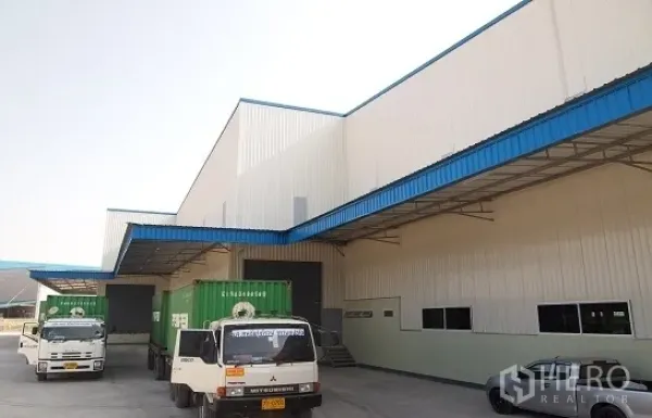 工厂 出租在Si Racha, Chon Buri - 带雨棚的装卸月台，集装箱卡车停靠在蓝白色现代工厂前。