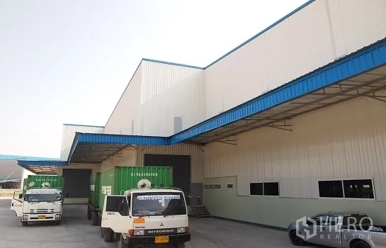 工厂 出租在Si Racha, Chon Buri - 带雨棚的装卸月台，集装箱卡车停靠在蓝白色现代工厂前。