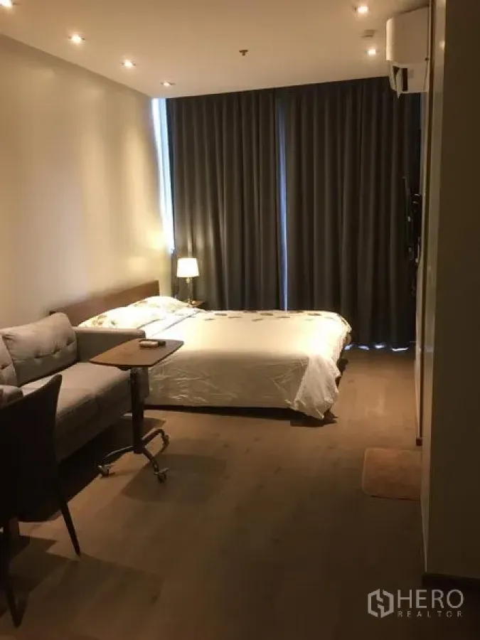 คอนโด ให้เช่าในKhlong Toei, Bangkok - โซนห้องนอนสไตล์สตูดิโอ เตียงคู่ ผ้าม่านทึบสีเทา โซฟาและโต๊ะข้าง ใต้ไฟดาวน์ไลท์