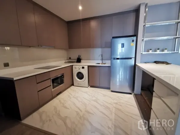 Condominium for rent or sale in Watthana, Bangkok - U-shaped kitchen with matte cabinetry, white countertops, induction hob, built-in microwave, washing machine, and stainless refrigerator on marble-look flooring.