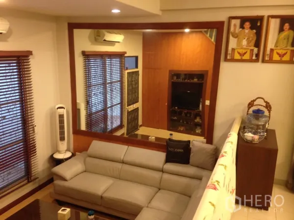 联排别墅 出租在Wang Thonglang, Bangkok - 客厅配灰色沙发、大型墙面镜与木质百叶窗，氛围温馨。