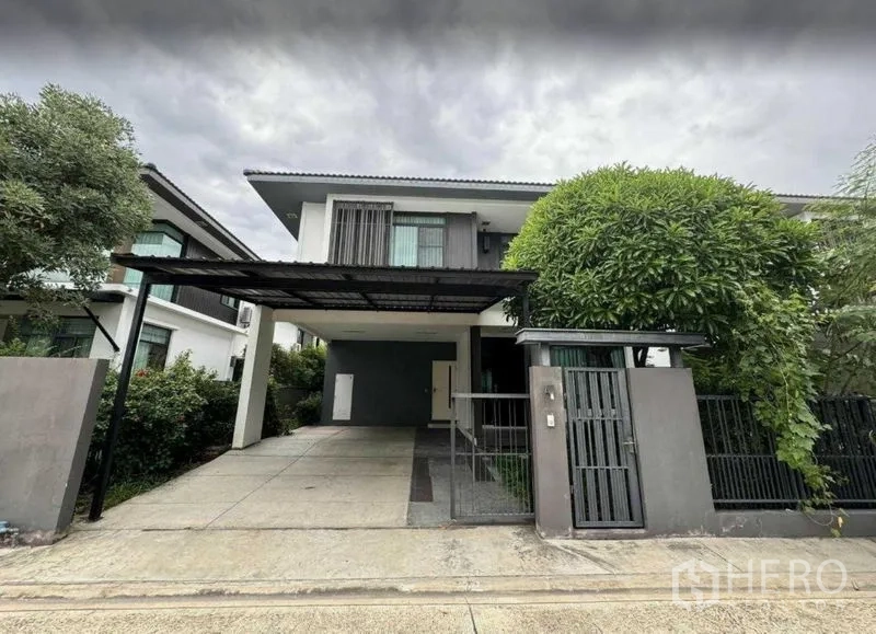 Detached House for rent in Samut Prakan - Wide driveway and metal gate leading to a two-storey home with balcony windows.