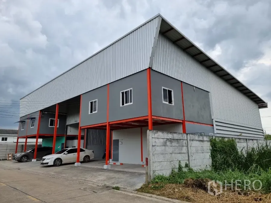 คลังสินค้า ให้เช่าในLam Luk Ka, Pathum Thani - ภายนอกโกดังสมัยใหม่ ผนังสีเทา เสาเหล็กสีแดง พร้อมที่จอดรถมีหลังคาในลำลูกกา