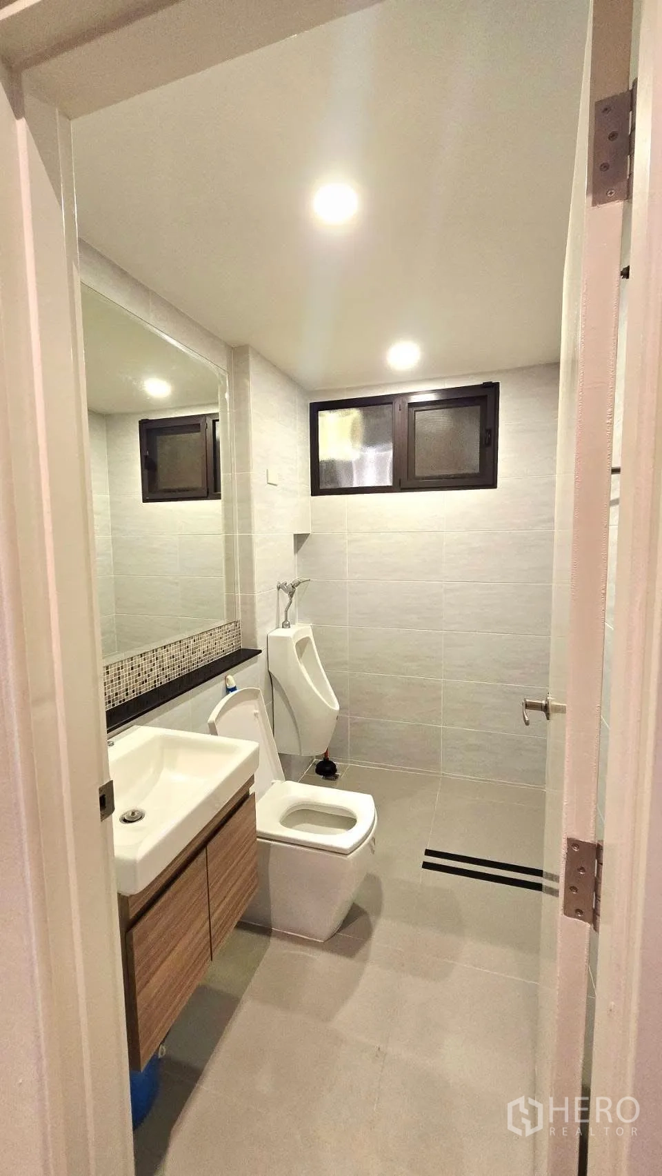 Detached House for rent in Prawet, Bangkok - Bathroom with sink vanity, toilet, urinal and grey wall tiles under recessed lighting.