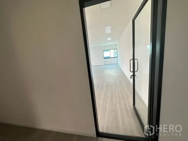 办公空间 出租 - 透过玻璃门可见明亮的开放式办公室，木纹地板、白色墙面与吸顶灯，尽头有窗户。